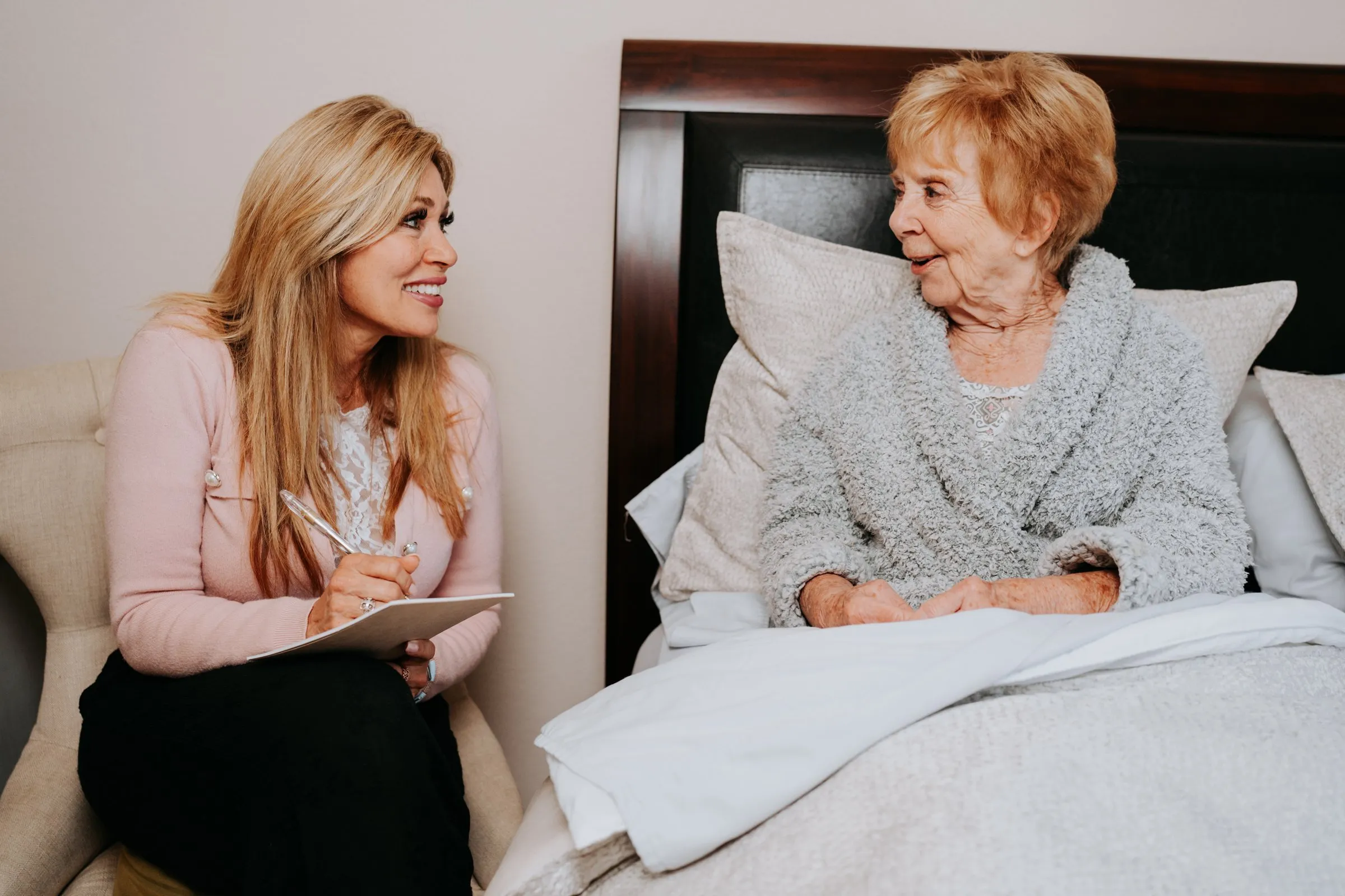 Michelle taking notes on a clipboard while an elderly client shares memories from her bed