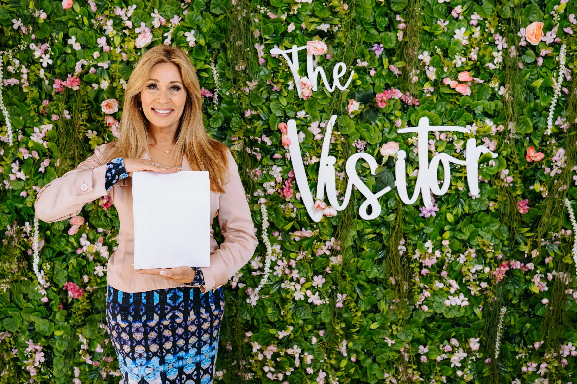 Michelle smiling and holding a written story in front of The Visitor flower wall